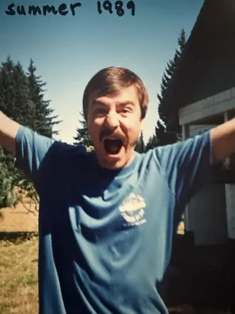 Robert in a blue t-shirt, arms thrown wide, in front of a cabin. Handwritten on the print: summer 1989.