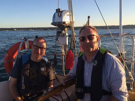 Robert and his nephew JP at the helm of a sailboat in life vests, city shoreline behind them.